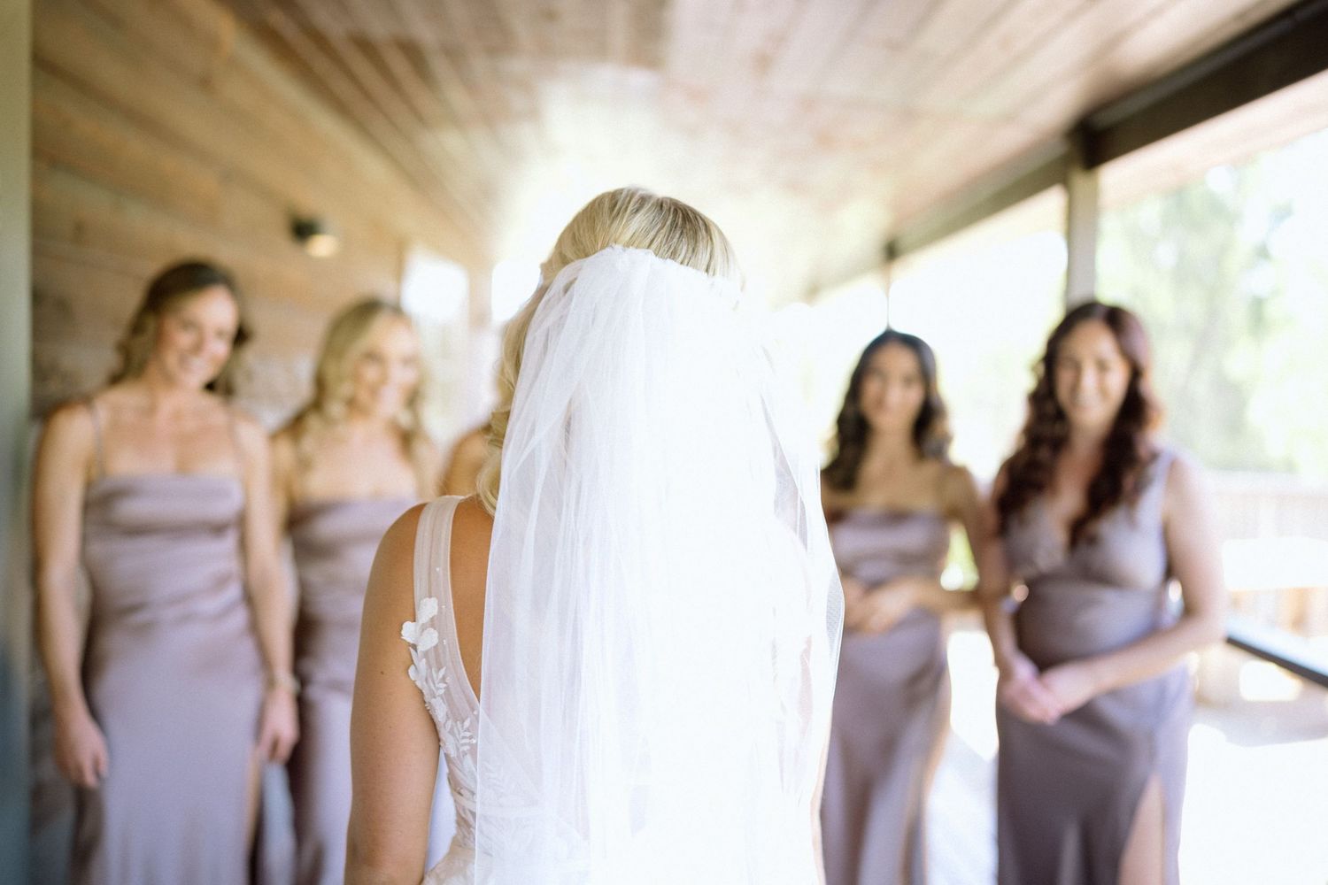 A bride in a veil stands with bridesmaids wearing lavender dresses in a covered walkway.