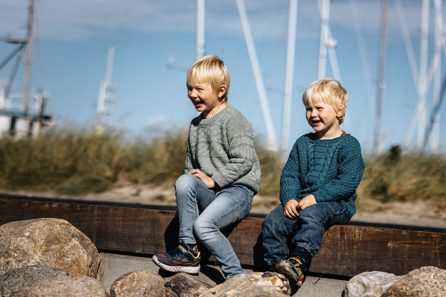 Two children in sweaters sit on coastal rocks near sailboat masts.