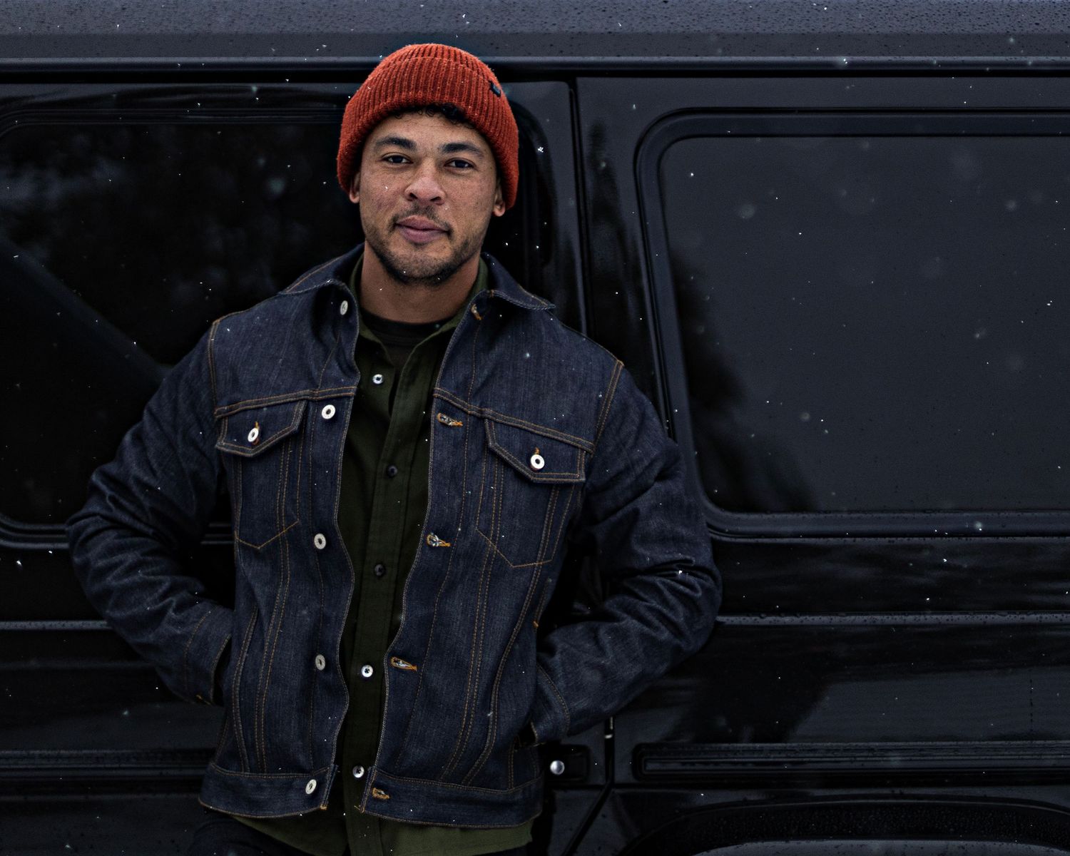 Someone wearing a red beanie and denim jacket standing by a black vehicle.