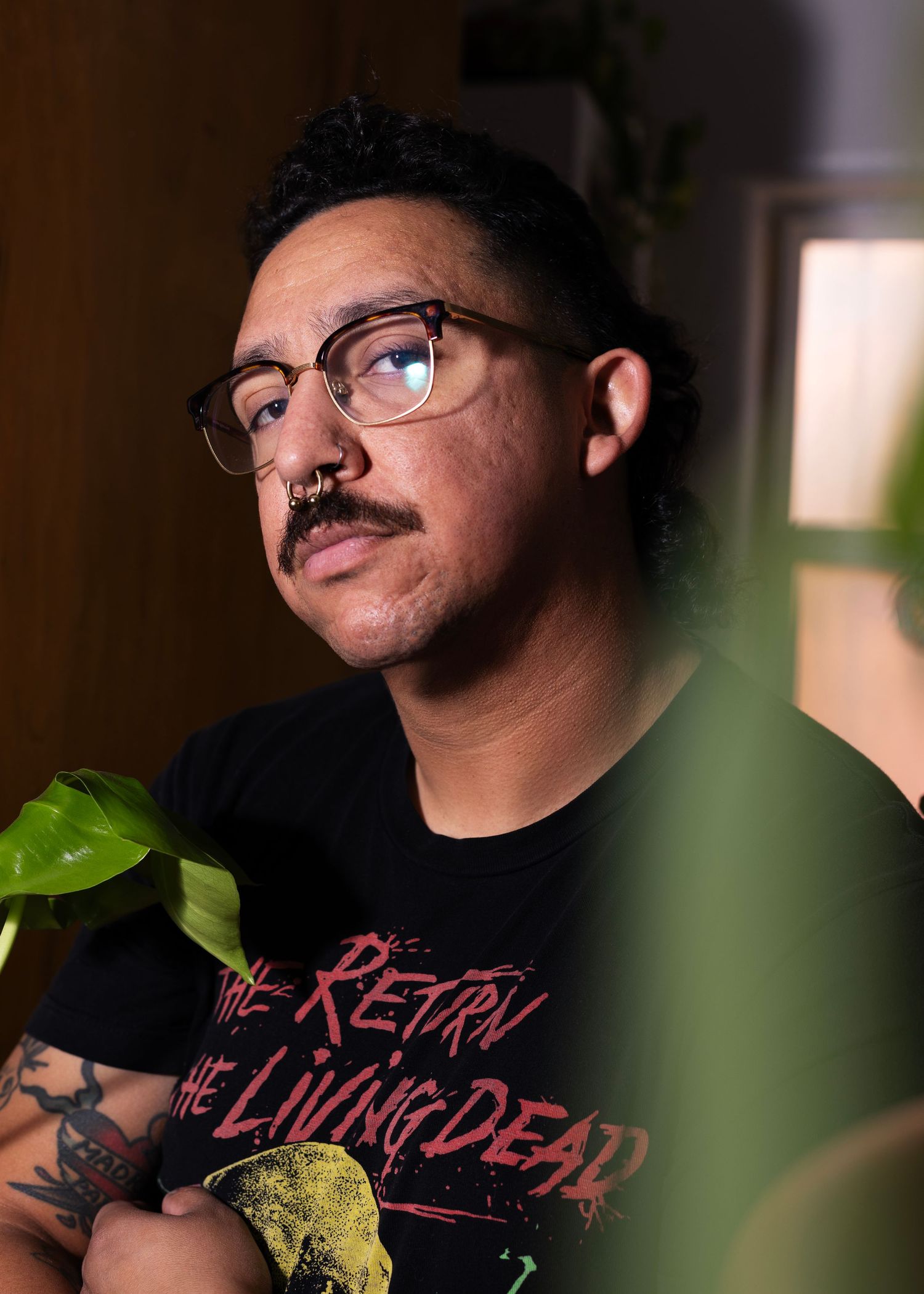 Portrait through plants of person wearing glasses and black t-shirt in warm lighting.