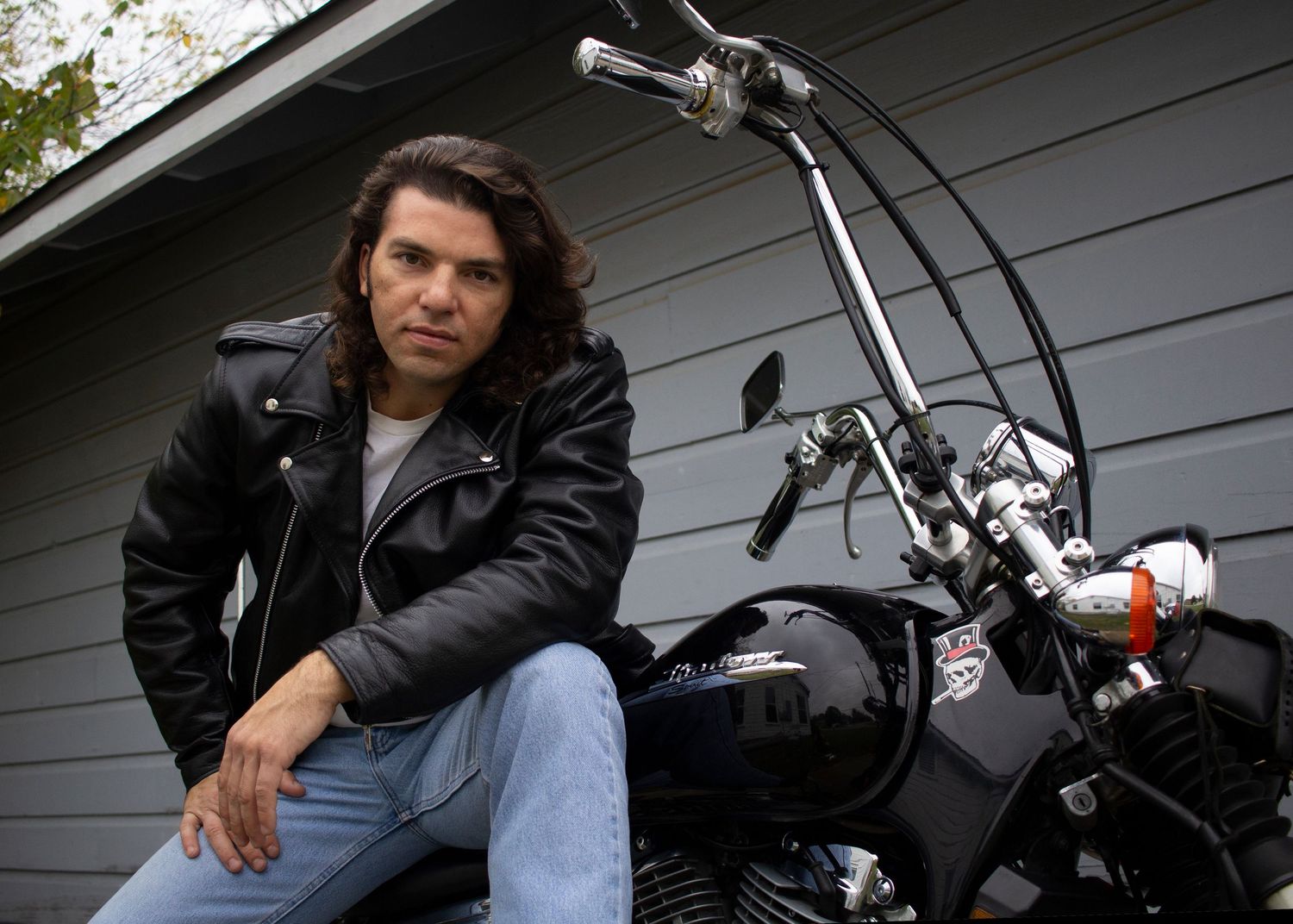 A black classic motorcycle parked by a gray house with someone wearing a leather jacket sitting next to it.