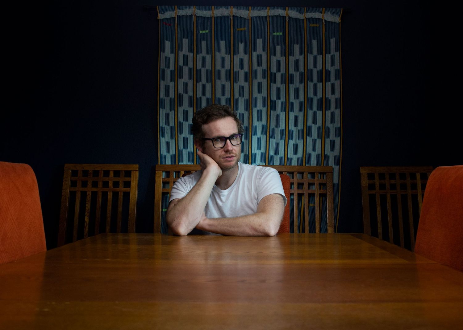A contemplative figure sits alone at a wooden table with orange chairs and a geometric patterned wall in the background.