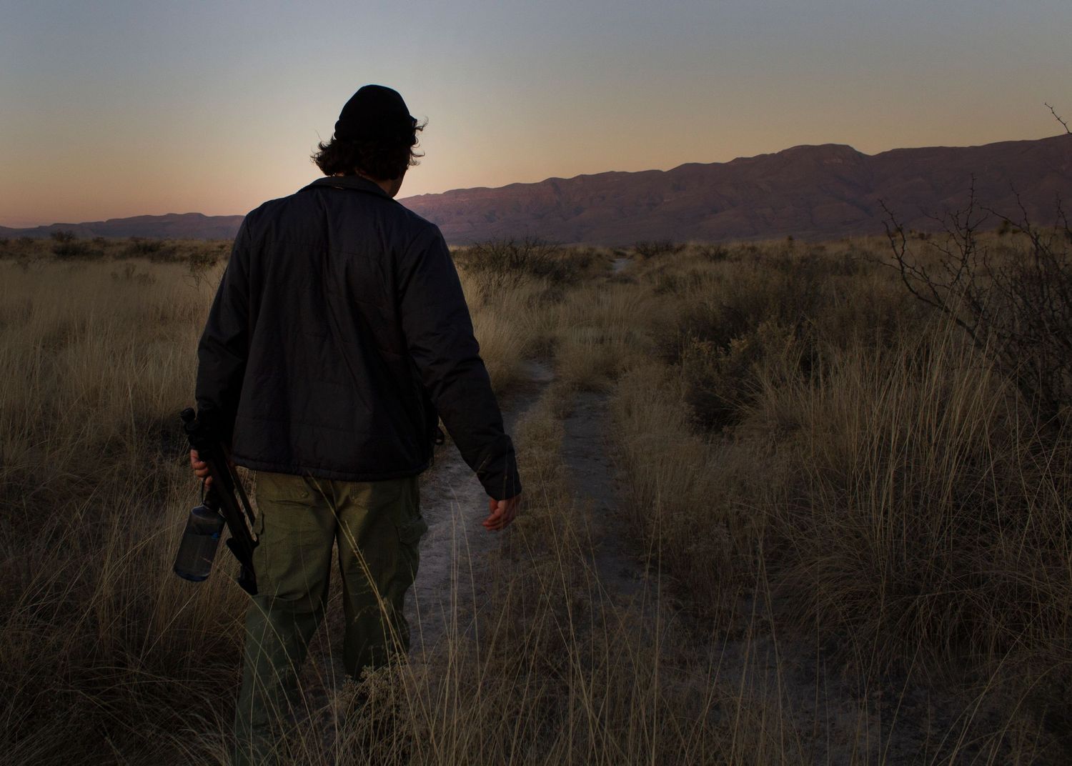 A silhouetted figure walks along a desert trail at dusk with mountains in the background.