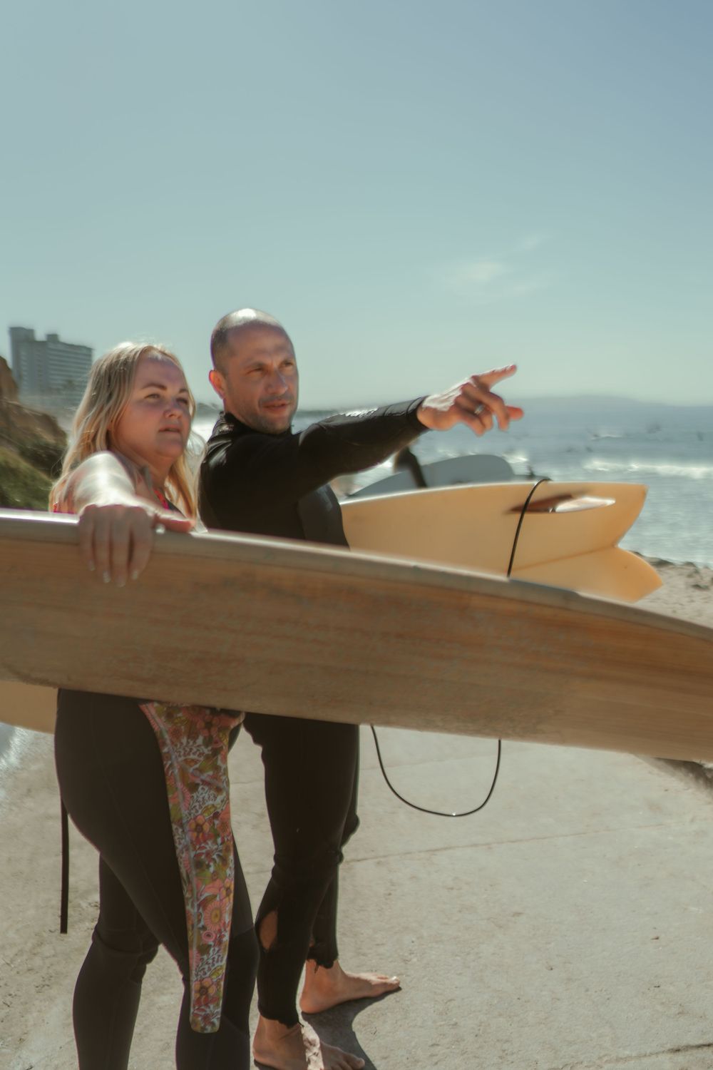 a couple standing on the beach in wetsuits with surfboards pointing toward the ocean