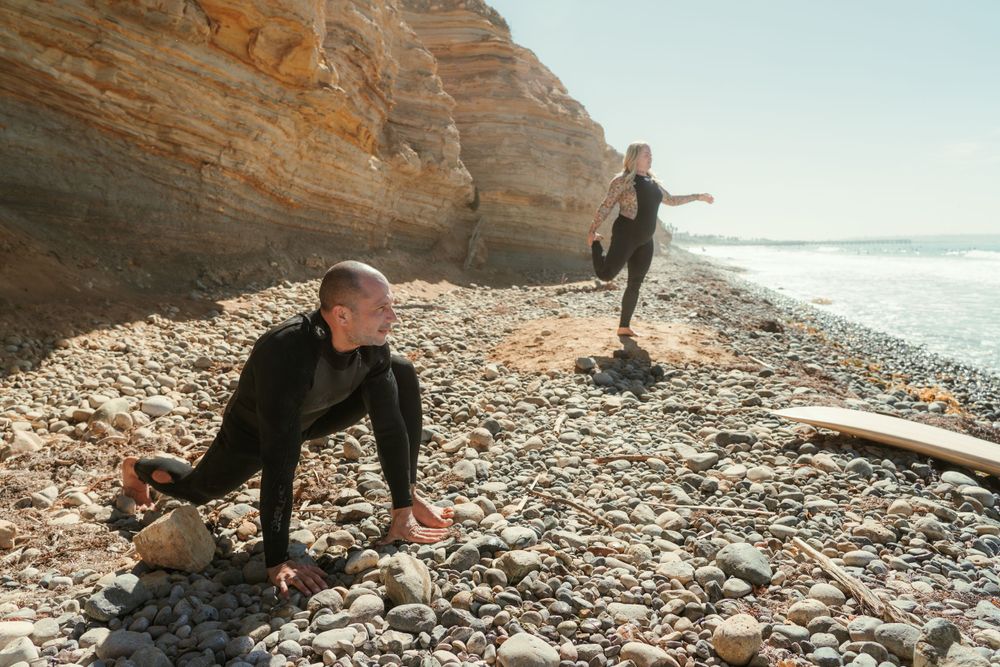 a couple stretching on the rocky ocean shoreline next to their surfboards