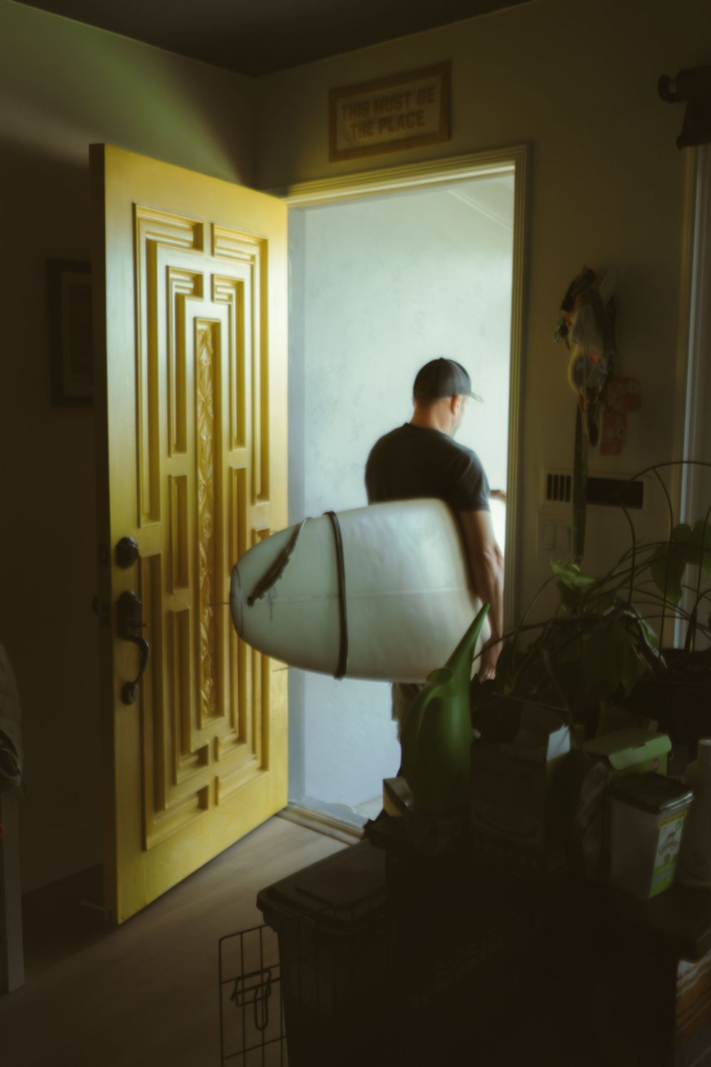 a man walking out a yellow front door carrying a surf board
