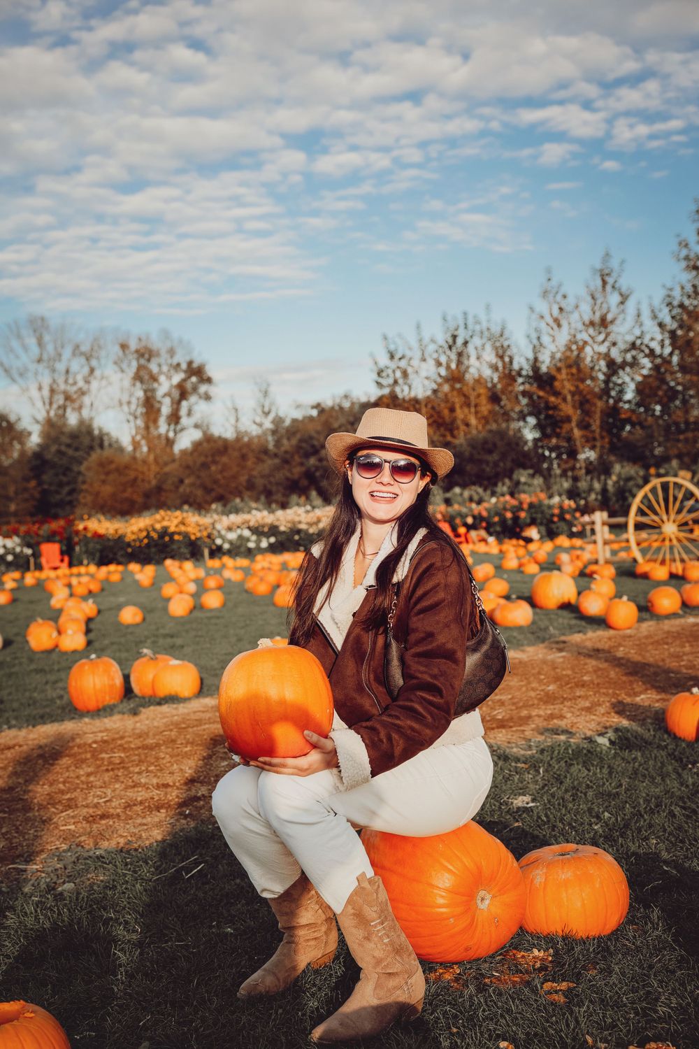 Woman in brown jacket and hat sitting on pumpkins at a rustic fall farm.