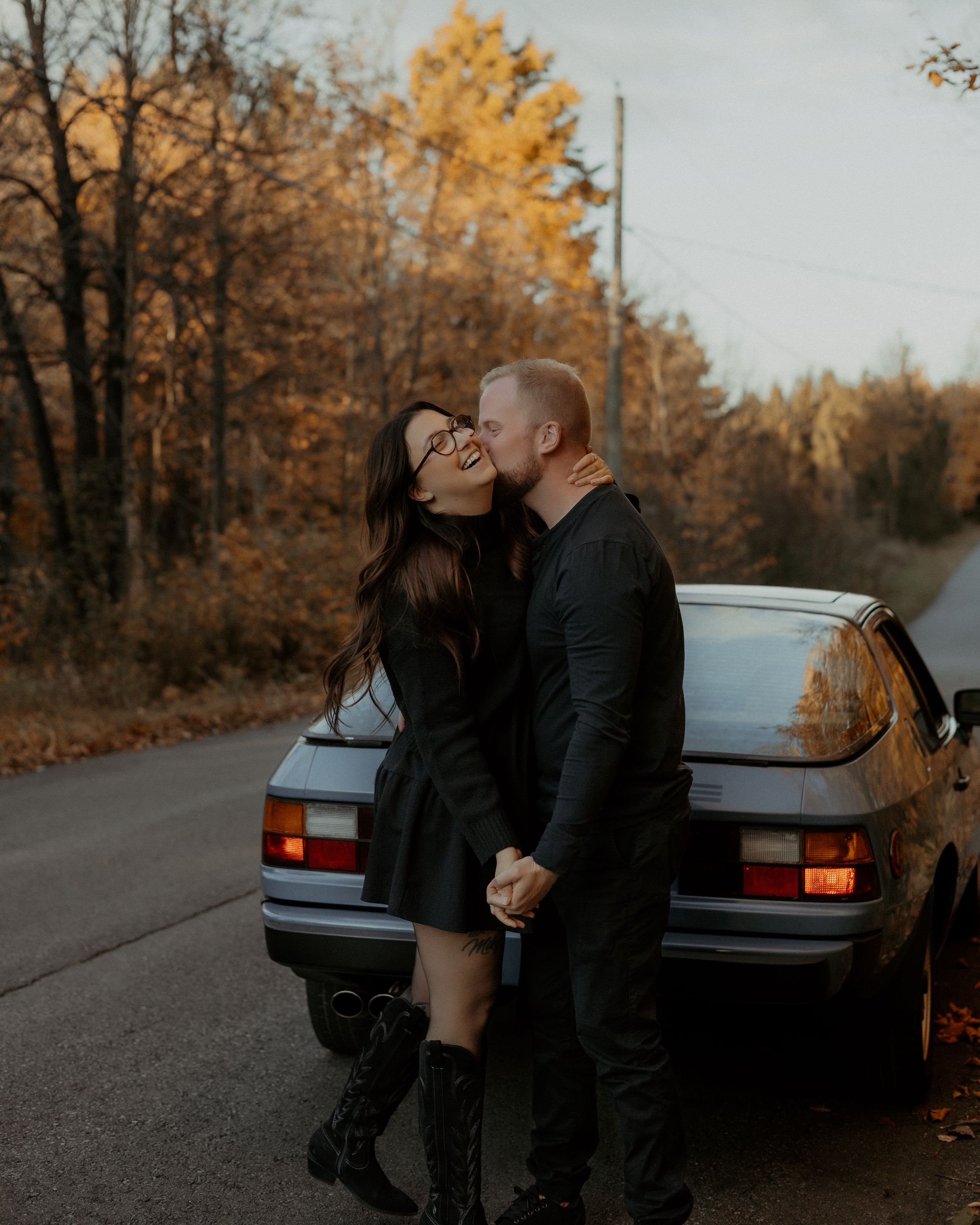 Autumn romance scene with couple by vintage car on tree-lined road.