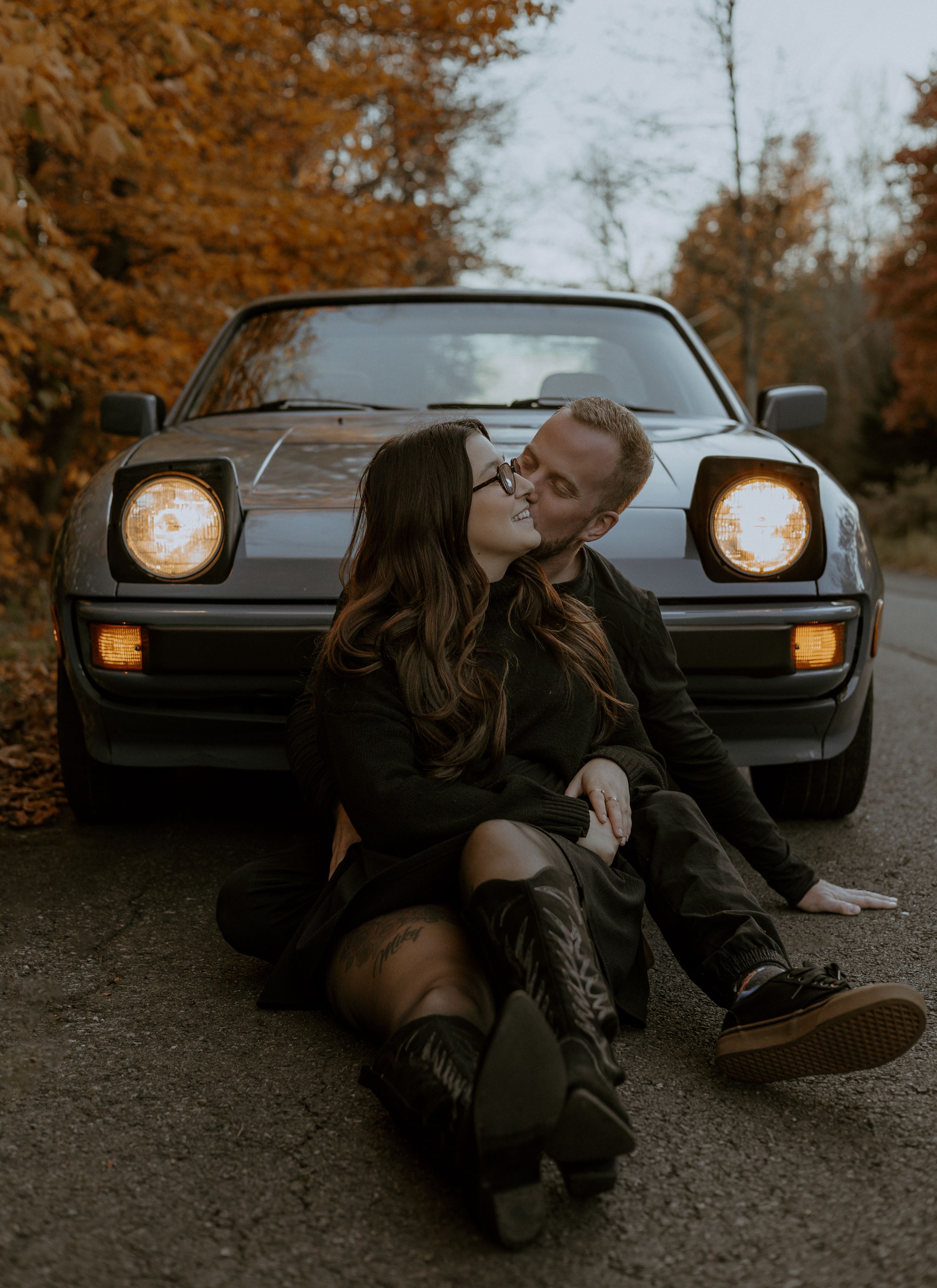 Intimate moment between couple sitting in front of classic car with illuminated headlights.