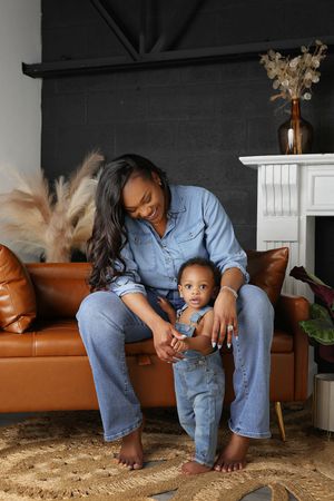Joyful mother in denim hugs her toddler in overalls on a leather couch near a white fireplace, with a rug and dried pampas grass.