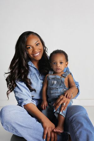 Smiling woman with long wavy hair in a denim outfit sits holding a barefoot toddler in denim overalls against a plain light studio backdrop.