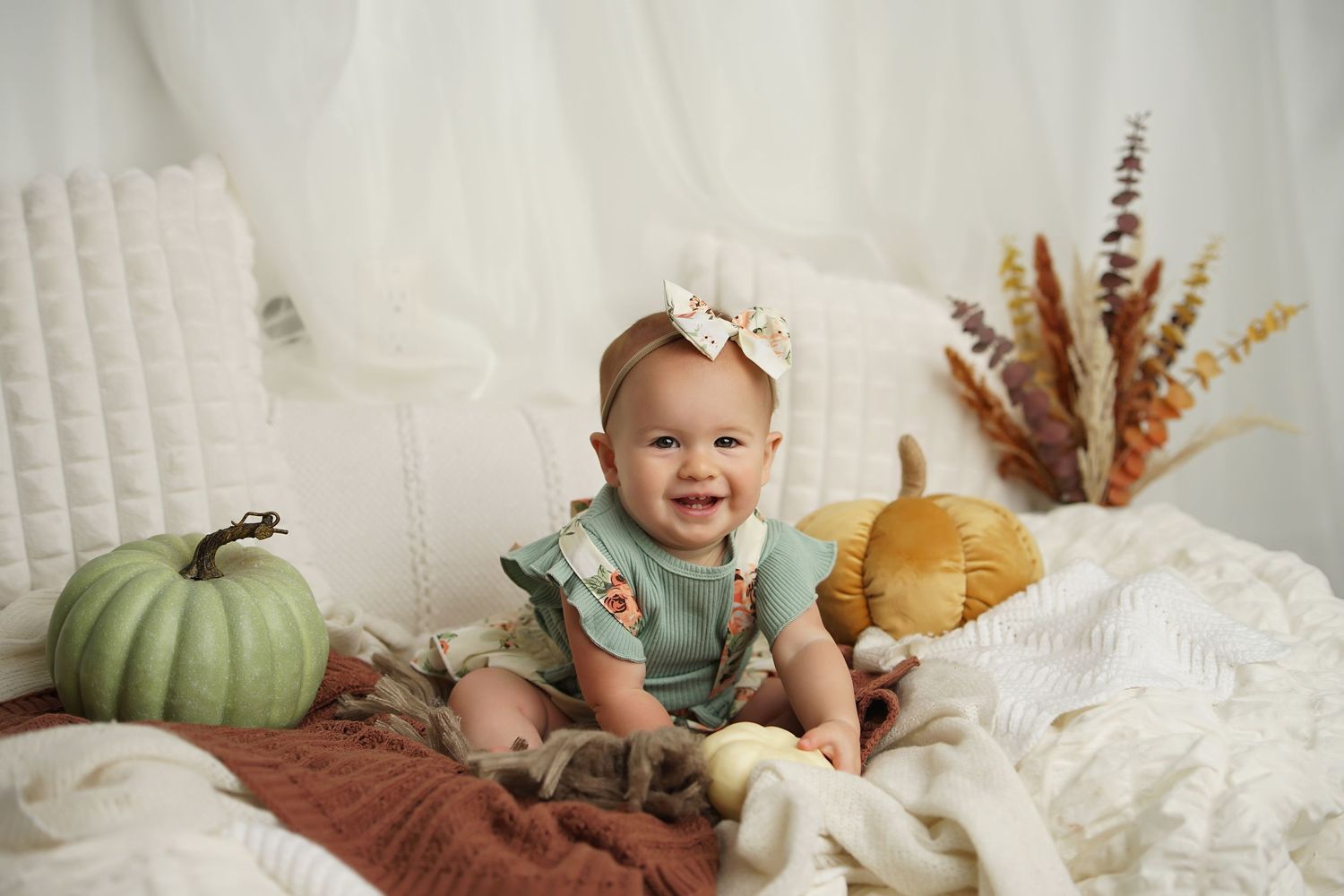 Smiling baby girl in a teal outfit and bow sits among green and yellow pumpkins on cozy neutral blankets for a fall photo.