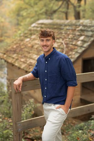 Smiling young man in a navy button-up shirt and light pants leans on a wooden fence near a rustic stone cabin with autumn foliage.