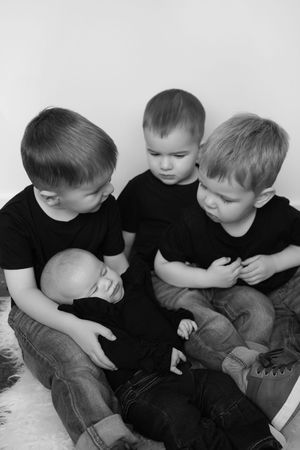 Black and white photo of three young boys in dark shirts and jeans sitting close together and gently cradling a sleeping newborn baby.