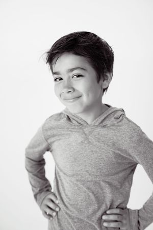 Black and white portrait of a smiling young boy with short dark hair, wearing a hooded long-sleeve shirt and posing with hands on hips.