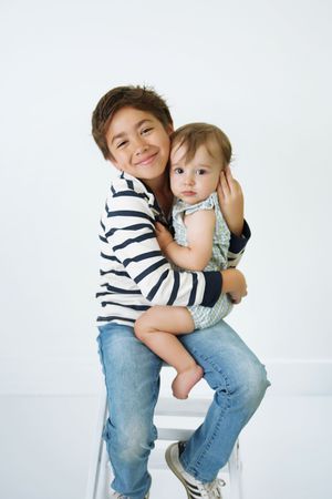 Smiling older boy in a striped shirt and jeans sits on a white stool, holding a baby in a light blue romper against a white background.