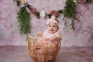 Baby girl in a pink outfit and floral headband sits in a woven rattan basket against a pink floral backdrop with hanging greenery.