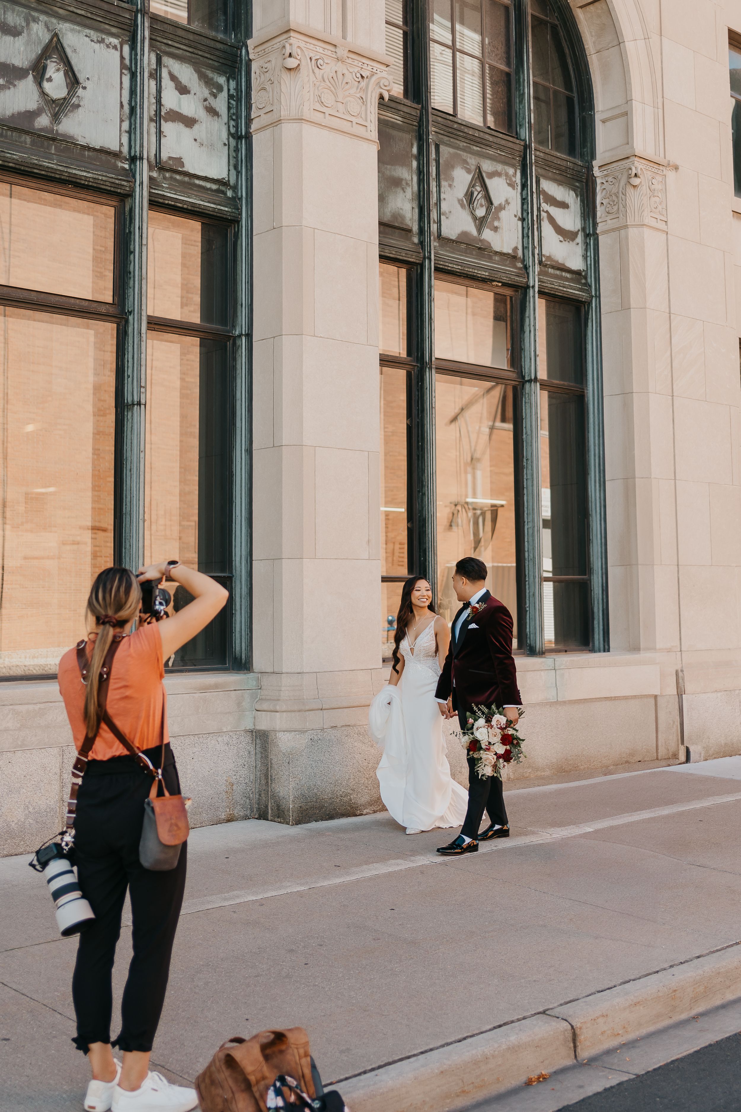 A photographer captures a couple posing against an ornate architectural backdrop with arched windows.