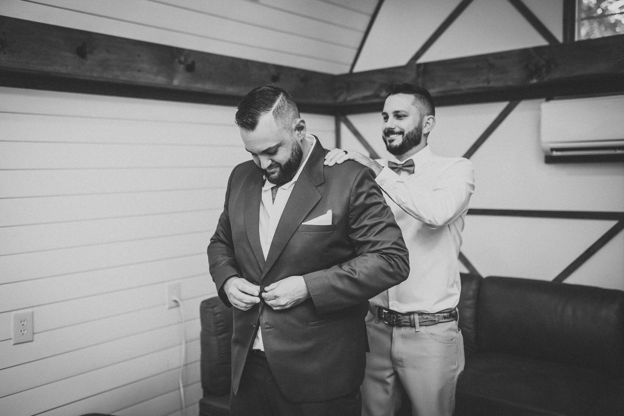 Groomsman helps groom prepare by adjusting his suit jacket in rustic indoor setting.