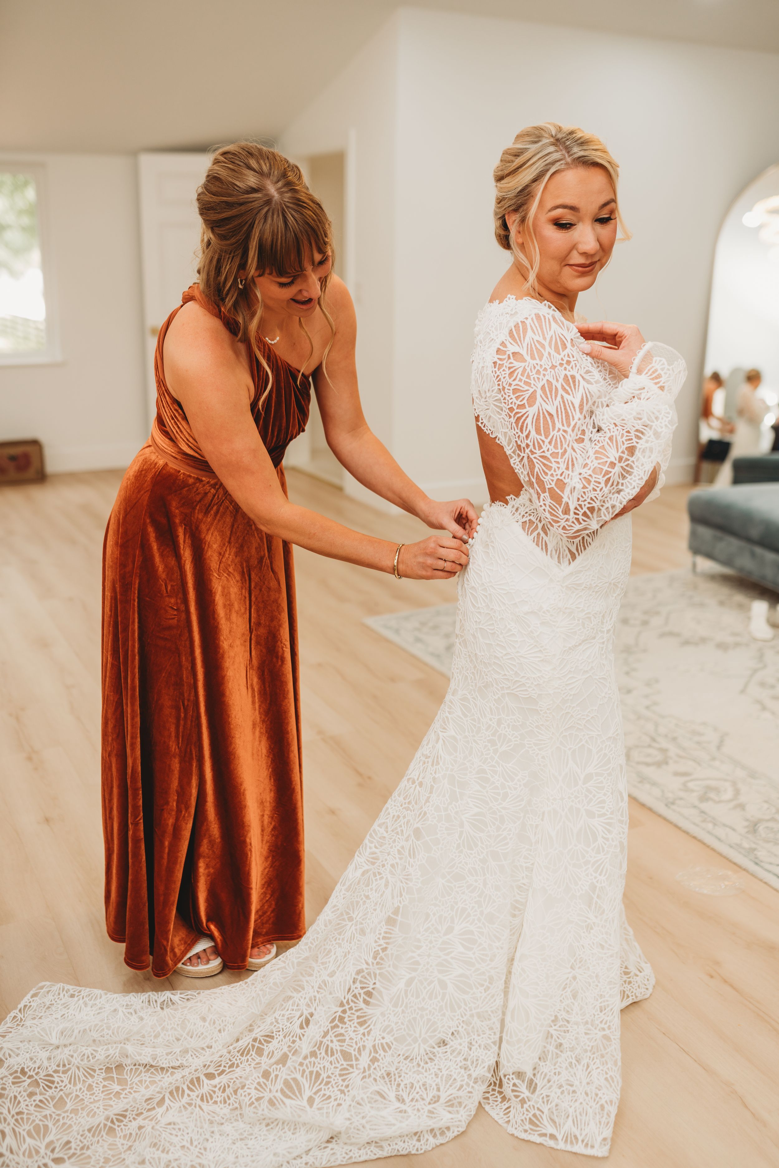 Bridesmaid in rust velvet dress helps bride with lace wedding gown fitting.