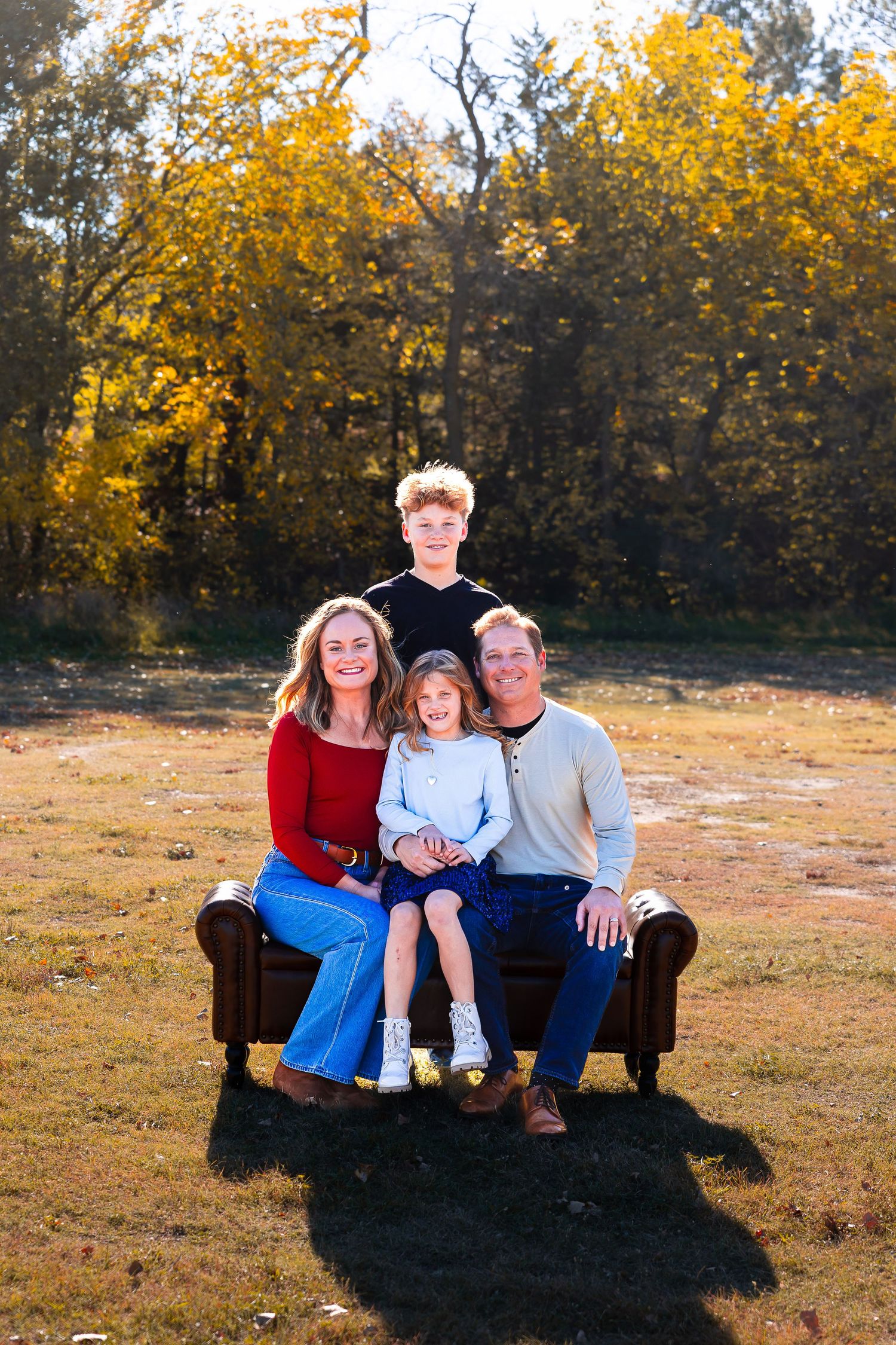 A group sits together on a brown couch outdoors with golden autumn foliage in the background during sunset.