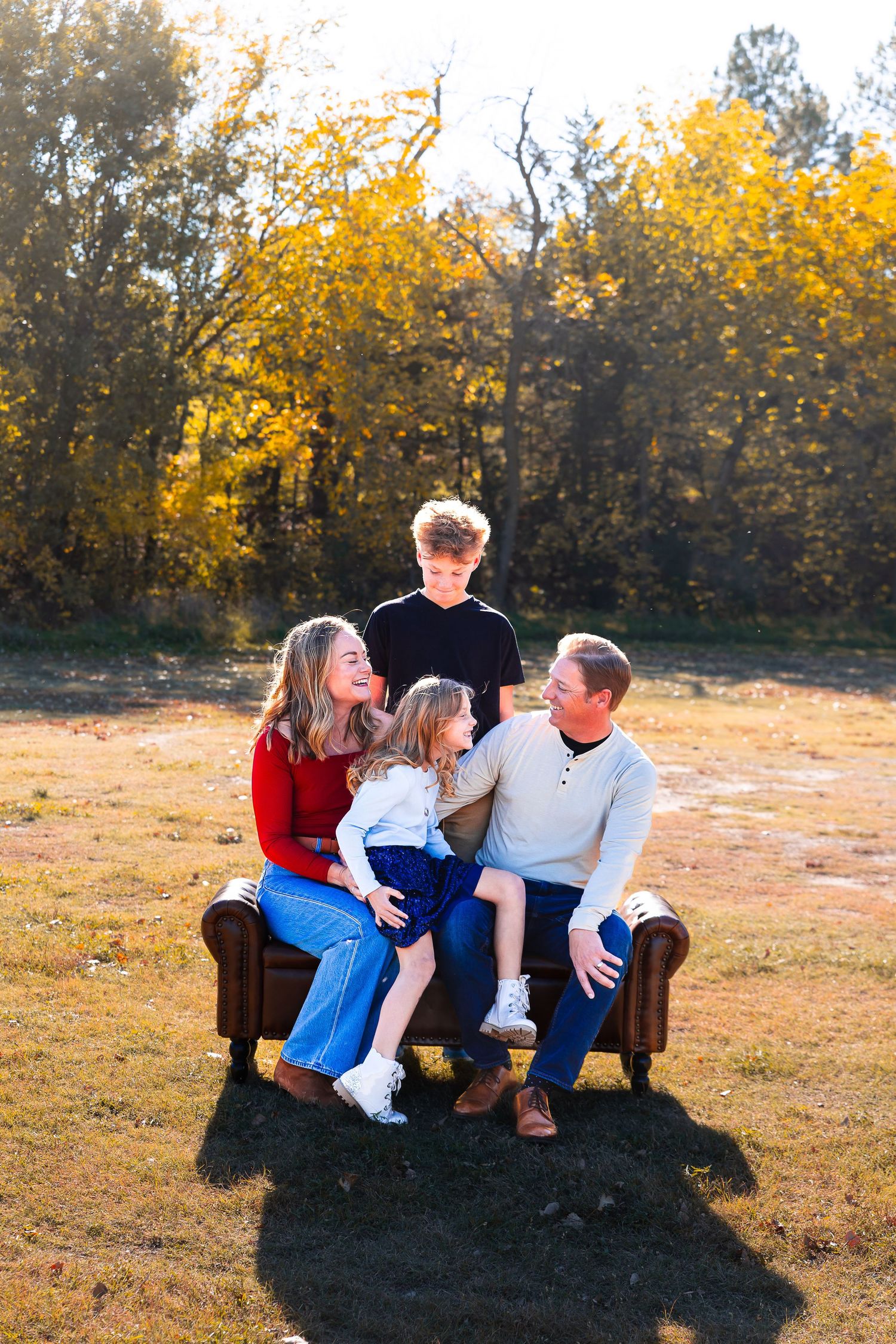 A group sits together on a brown leather couch outdoors during golden hour with autumn trees in the background.