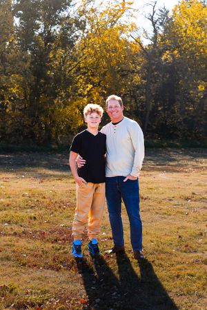Two people stand together in a sunny autumn field with golden foliage and long shadows cast behind them.