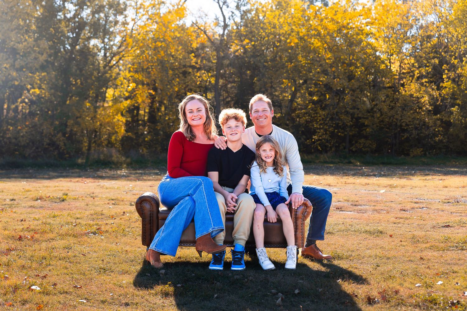 A family of four sits together on a brown couch outdoors with golden autumn foliage in the background.