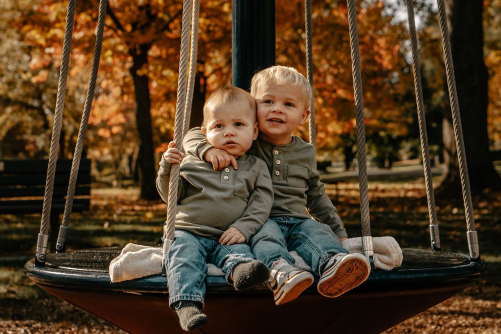 Two young children sitting together on round swing during fall with orange leaves in background.