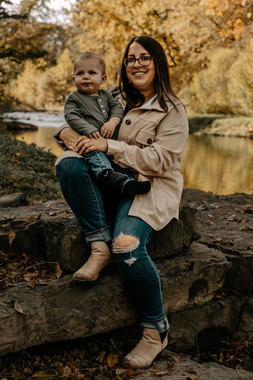 Parent and child sitting on rocky ledge by water during autumn with golden foliage in background.