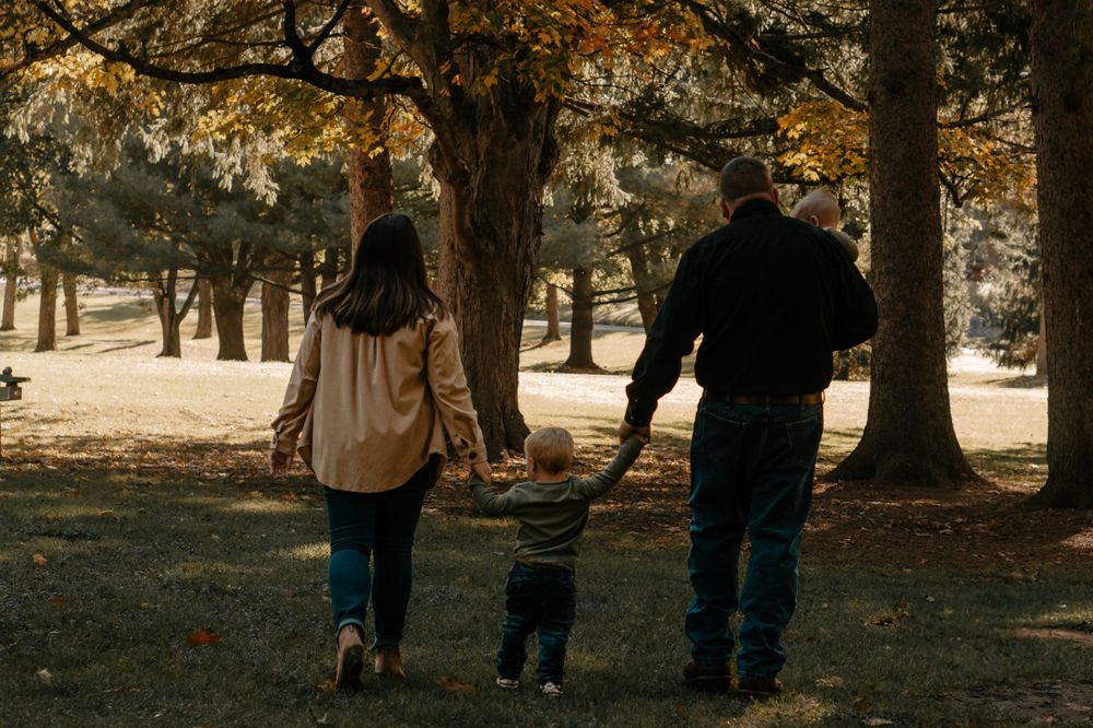 Family holding hands while walking through park with autumn trees casting long shadows.