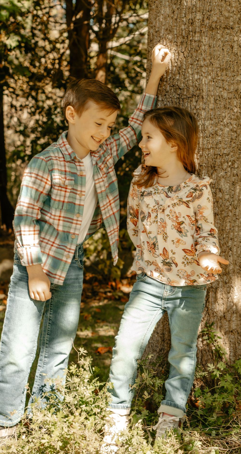 Two children in casual wear playing and laughing together by tree trunk in sunlight.