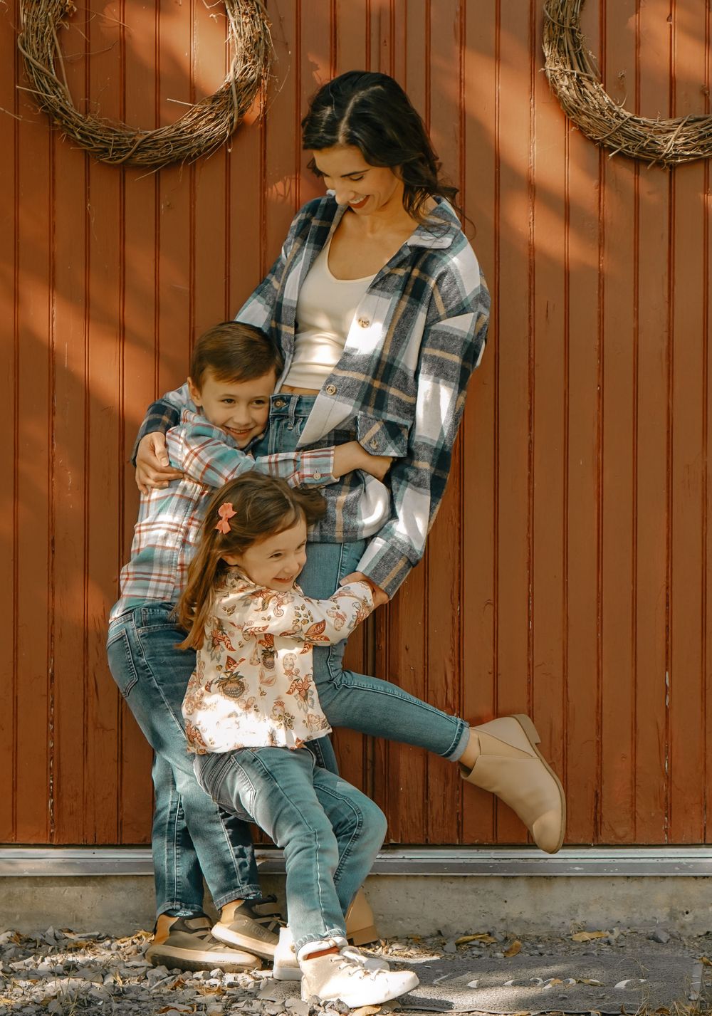 Family in coordinating plaid and floral outfits leaning against rustic wooden wall with decorative wreaths.