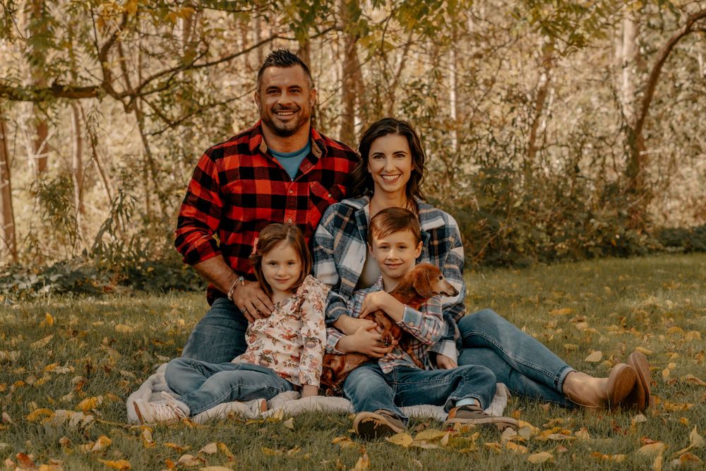 Family of four in coordinating plaid shirts sitting together on grass during autumn photoshoot.