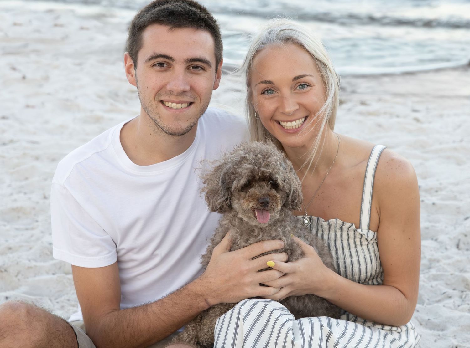 Young couple smiles with gray poodle on sandy beach.