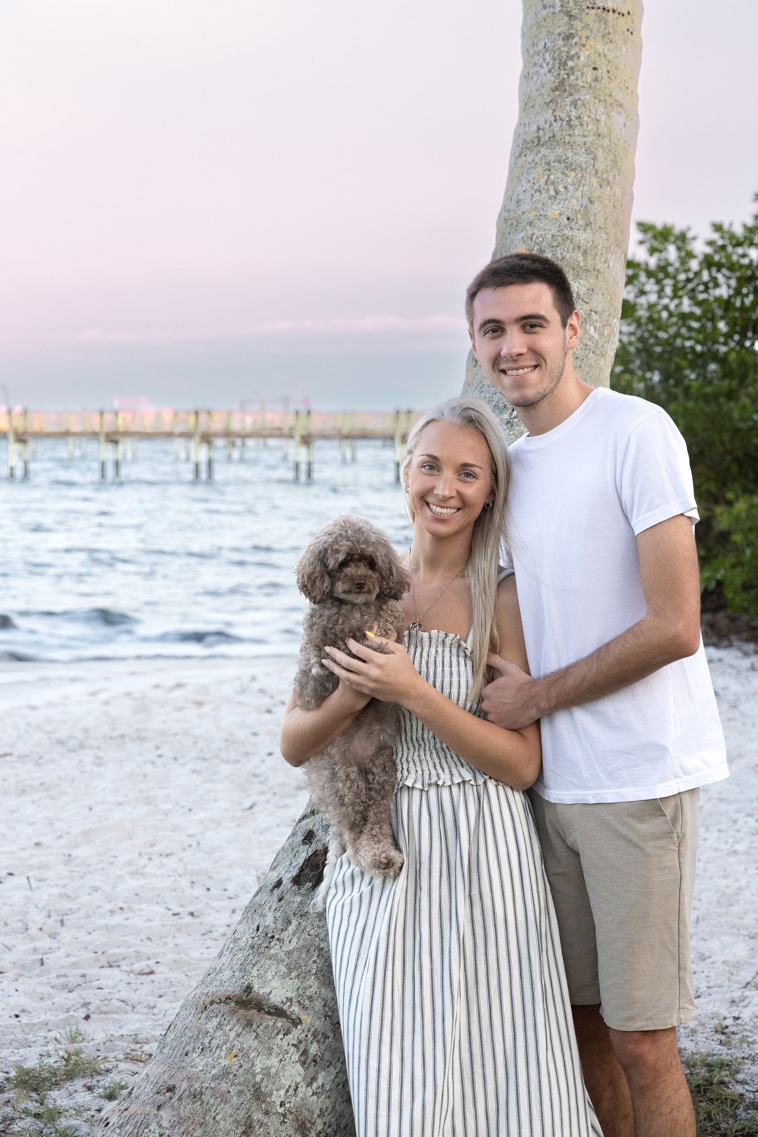 Smiling couple holds fluffy dog near palm tree on tropical beach.