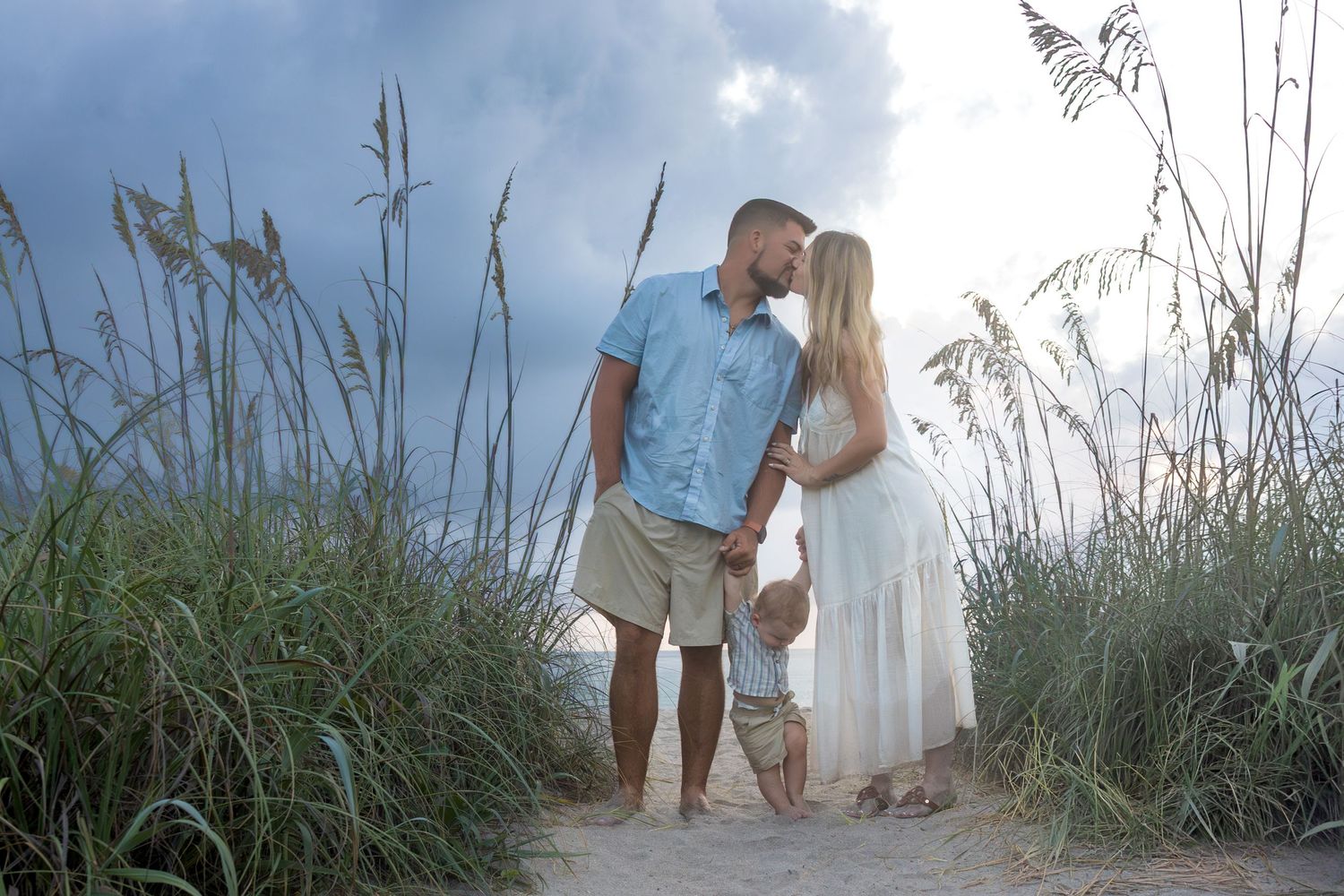 Couple sharing a romantic kiss on a sandy beach path surrounded by tall sea oats under a dramatic cloudy sky.