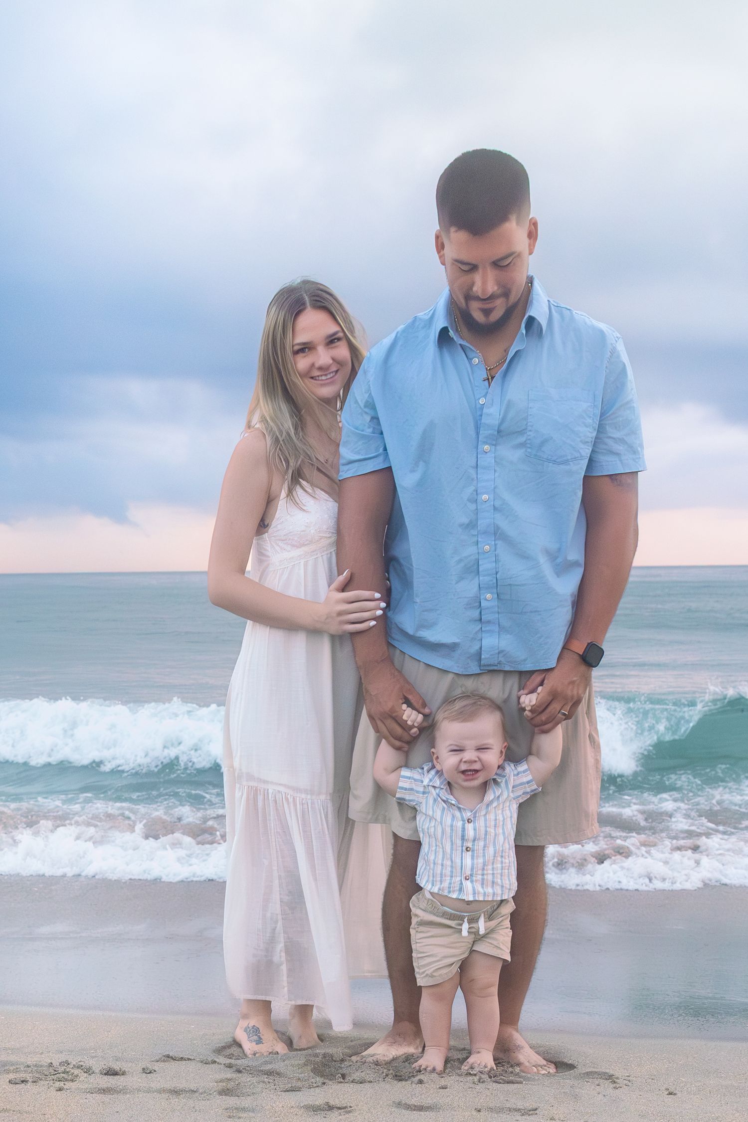 Happy family of three poses on a sandy beach at sunset, parents holding toddler son's hands near ocean waves.