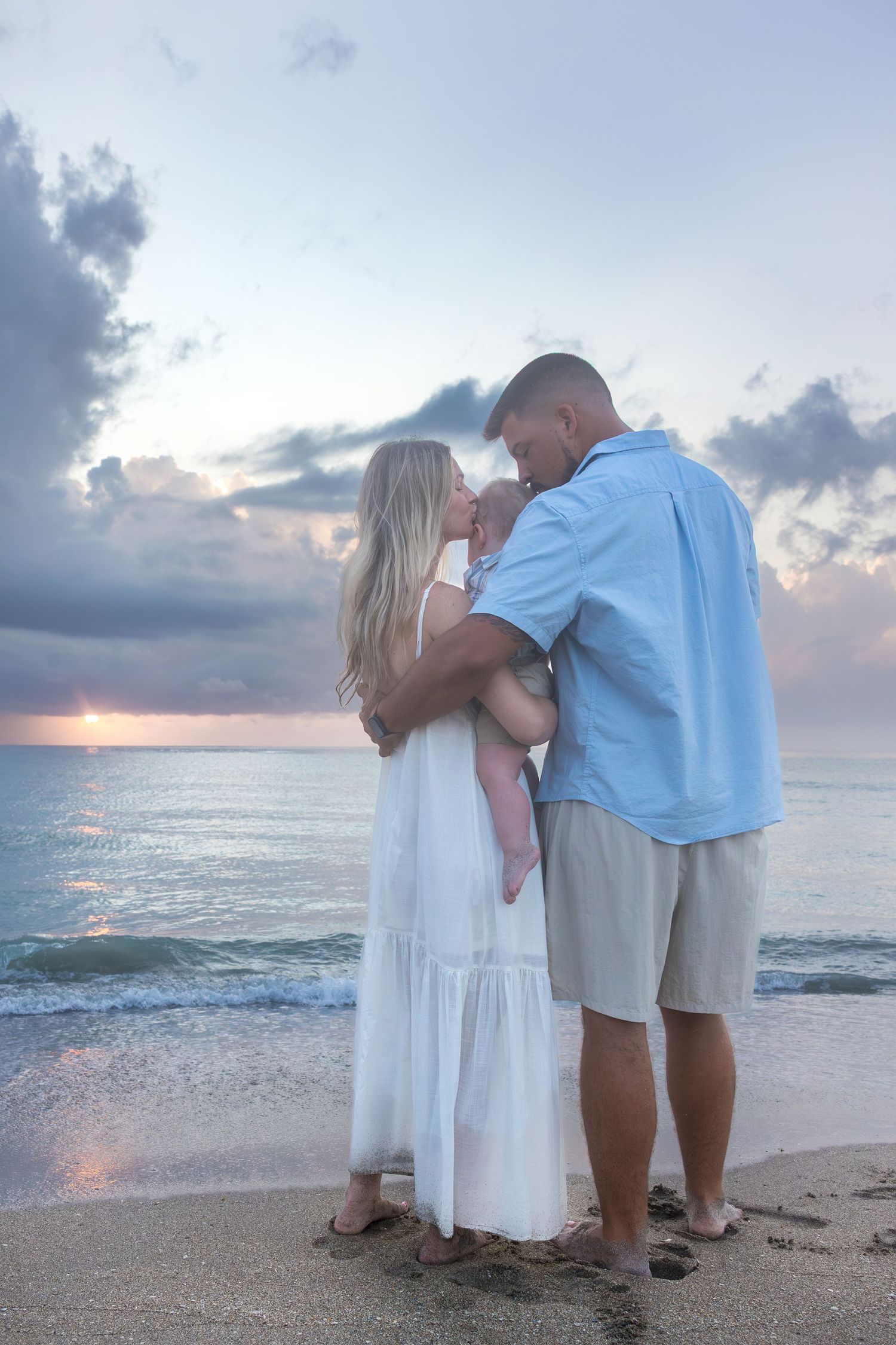 Couple embracing on a beach at sunset, woman in white dress, man in blue shirt, holding a baby by the ocean.