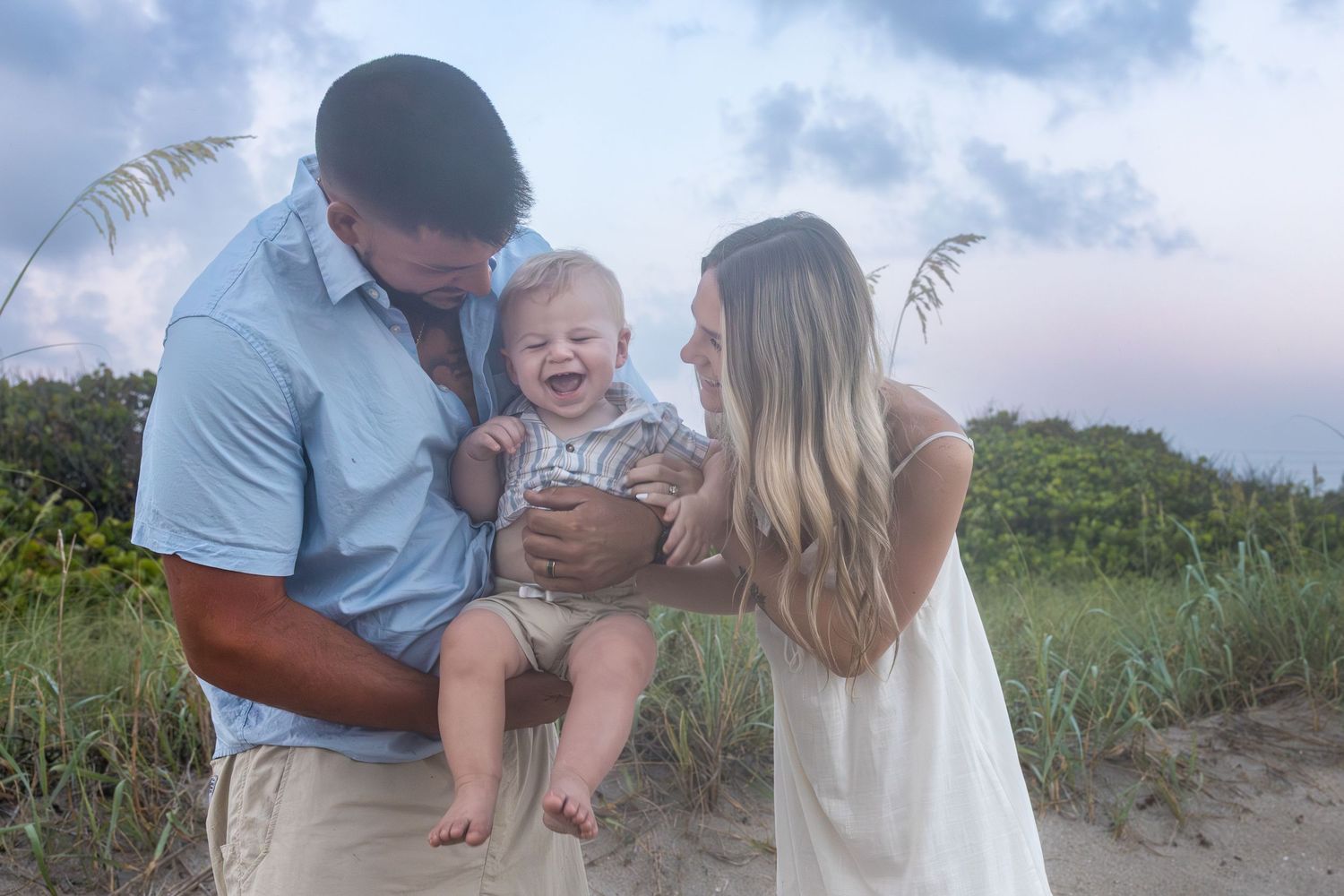 Happy family with parents holding laughing baby on sandy beach at sunset with lush greenery backdrop.