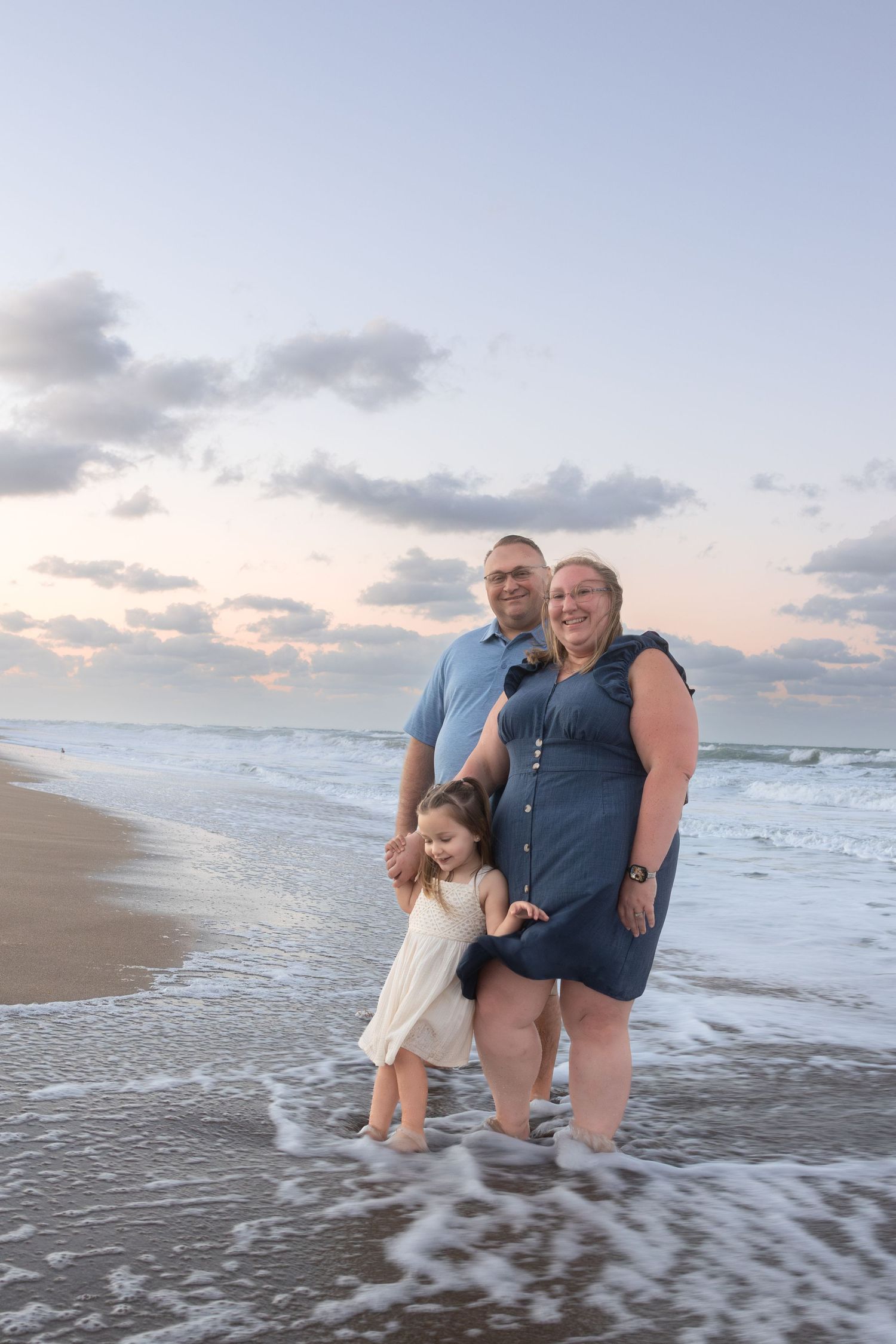 Family of three stands in ocean waves at sunset on beach.