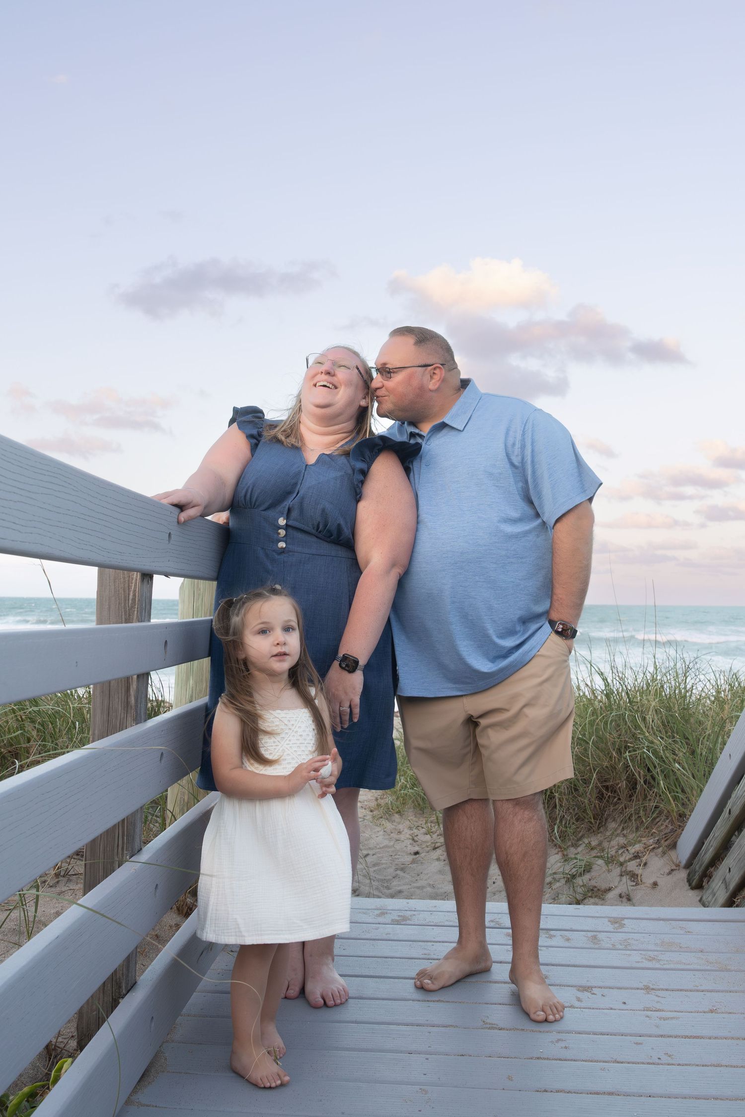 Couple shares kiss on boardwalk while daughter stands nearby.