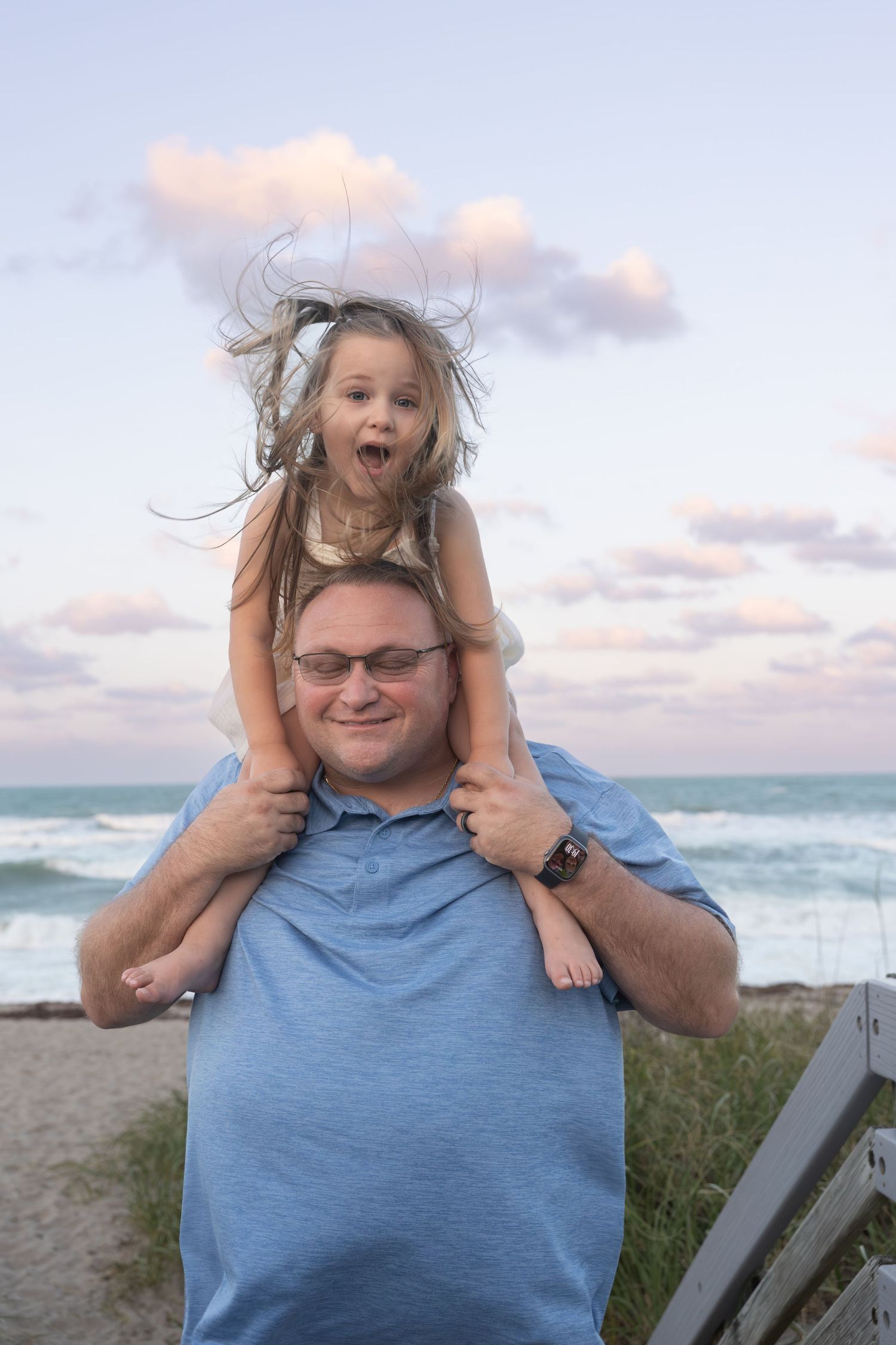 Smiling father carries laughing girl on shoulders at beach sunset.