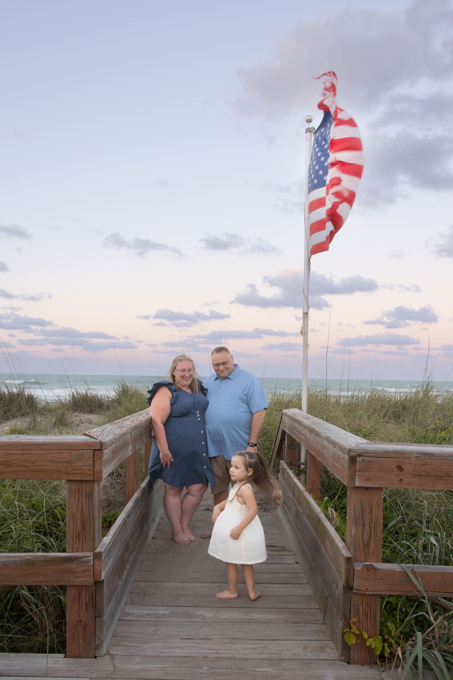 Family poses on boardwalk with American flag at beach sunset.