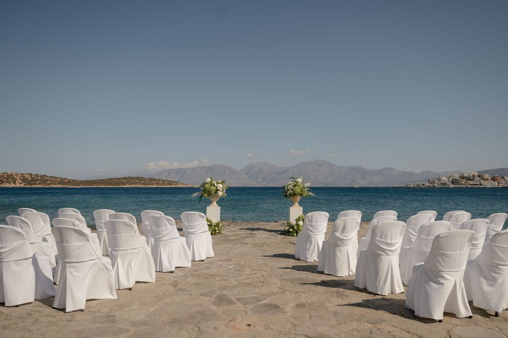 White chairs set up for beach wedding ceremony with ocean and mountains in background on sunny day.