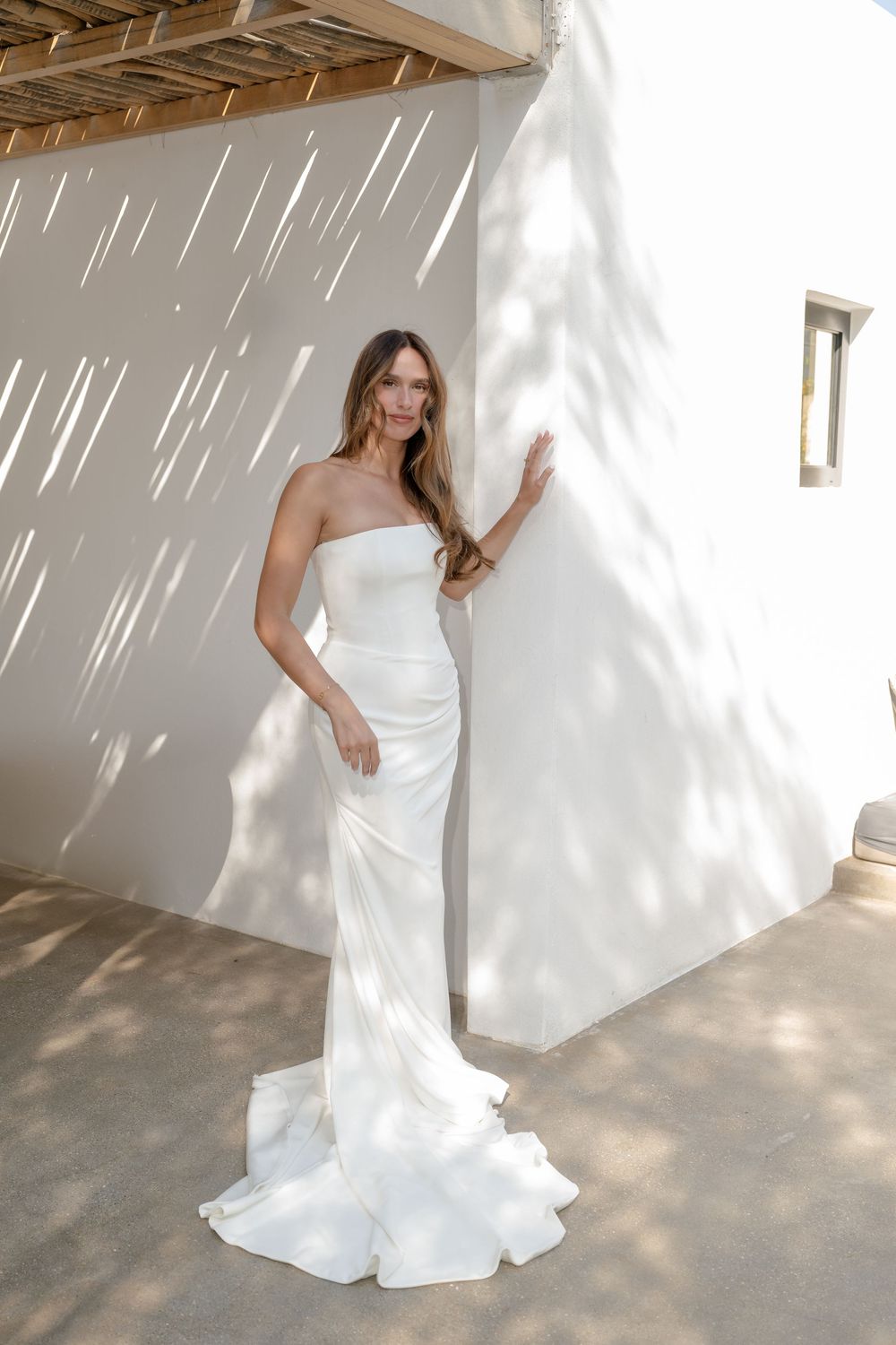 A bride in a white strapless wedding gown poses against a sunlit white wall creating dramatic shadows.
