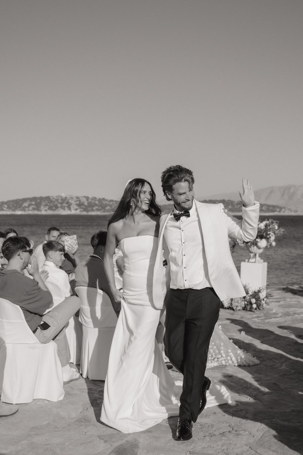 Wedding guests in formal attire sit in white-covered chairs arranged on a beach with ocean views.