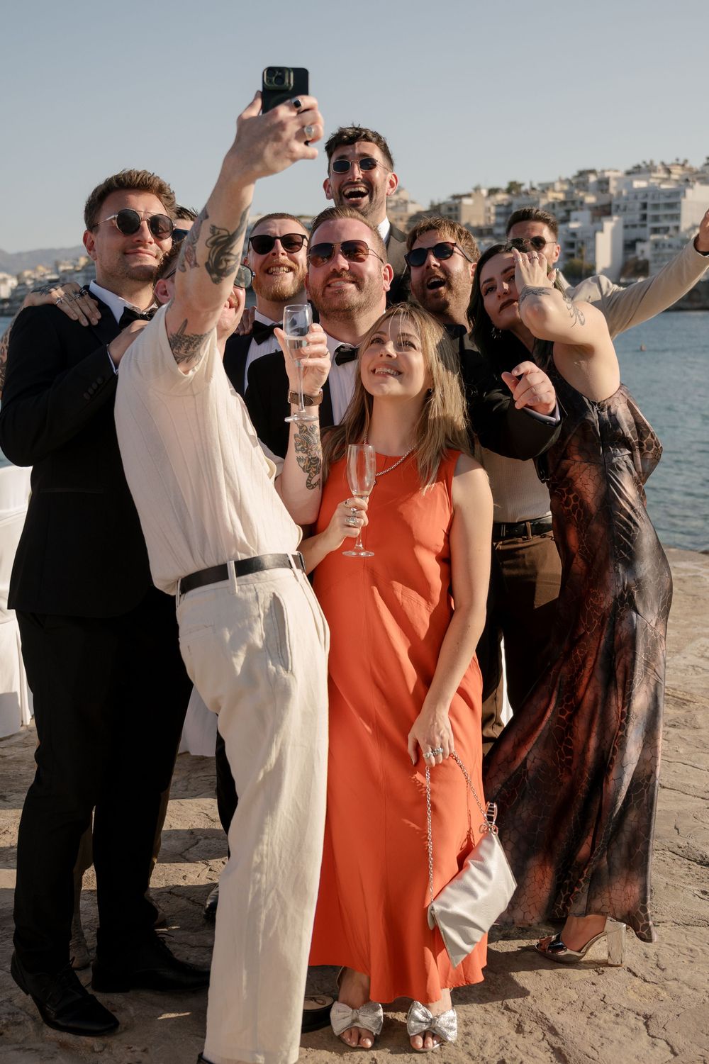Wedding party celebrates on a beach with guests in orange and brown dresses posing for photos.