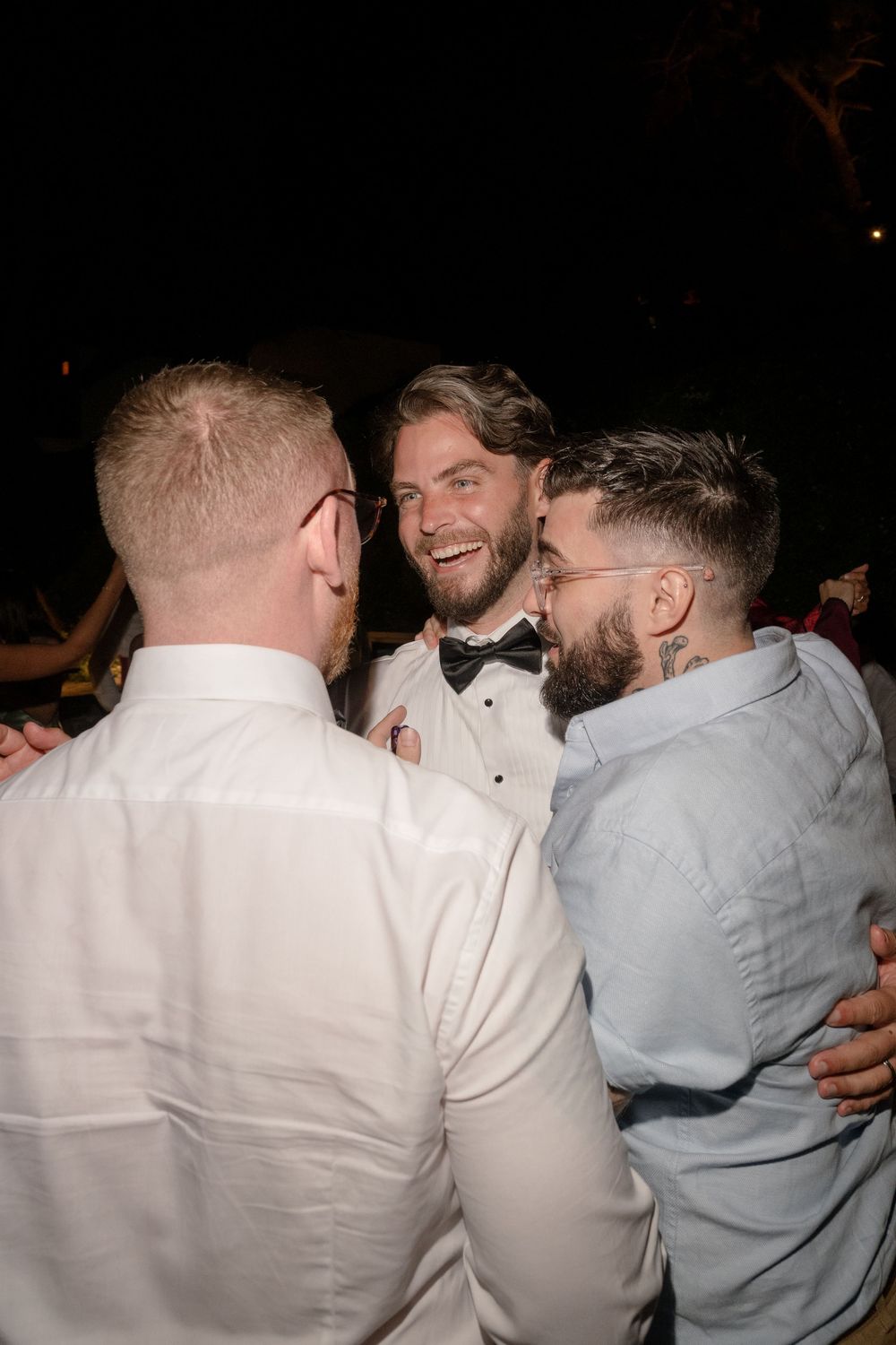Group of wedding guests in white and light colored formal attire laughing and embracing at a celebration.
