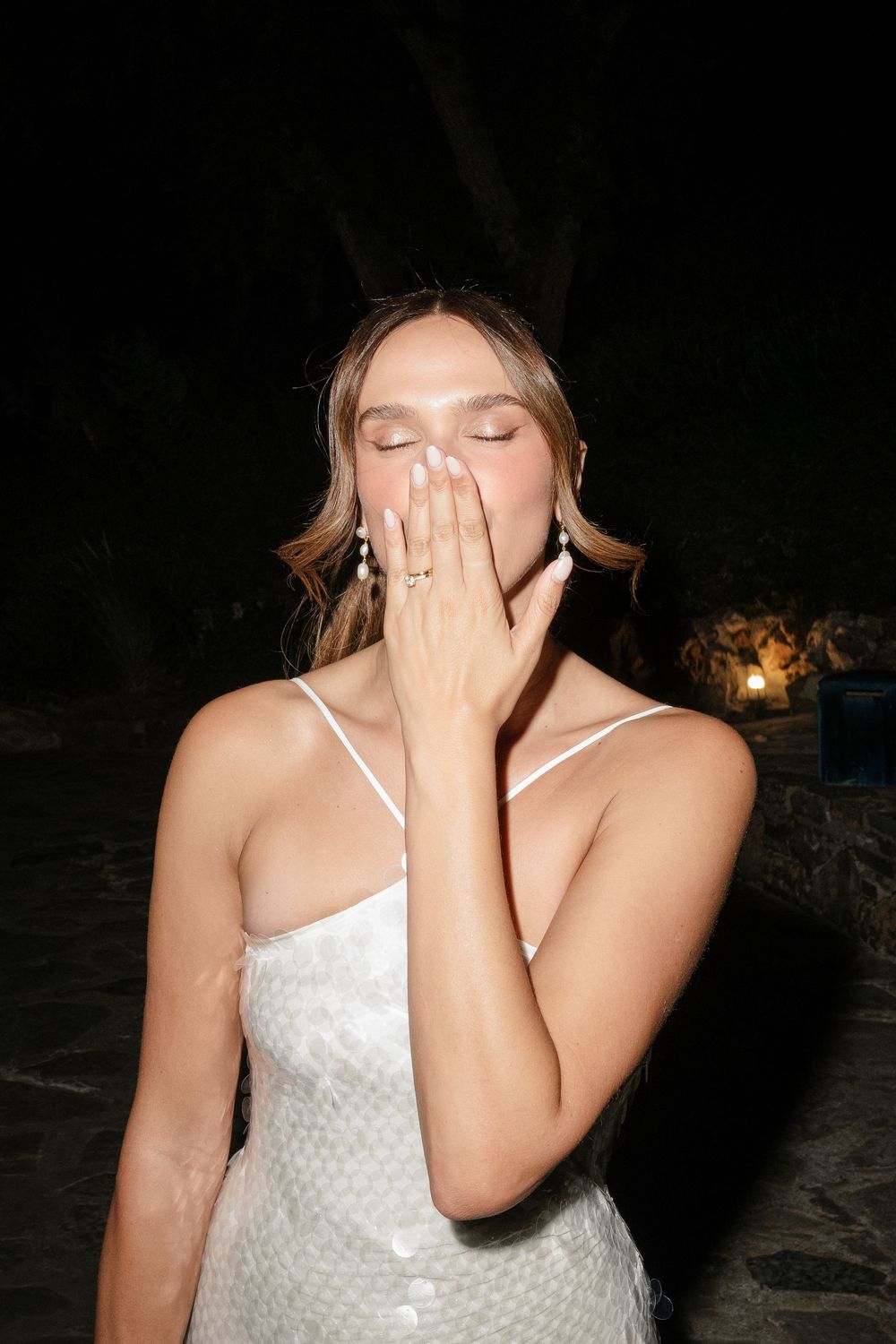 Woman in white dress covering her face while laughing during a nighttime celebration.